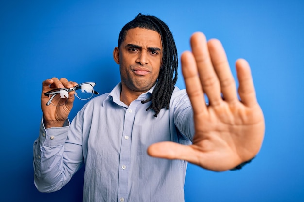 Joven afroamericano con rastas sosteniendo anteojos sobre fondo azul con la mano abierta haciendo señas de alto con gesto de defensa de expresión seria y confiada