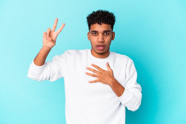 Joven afroamericano rizado aislado en azul tomando un juramento, poniendo la mano en el pecho.