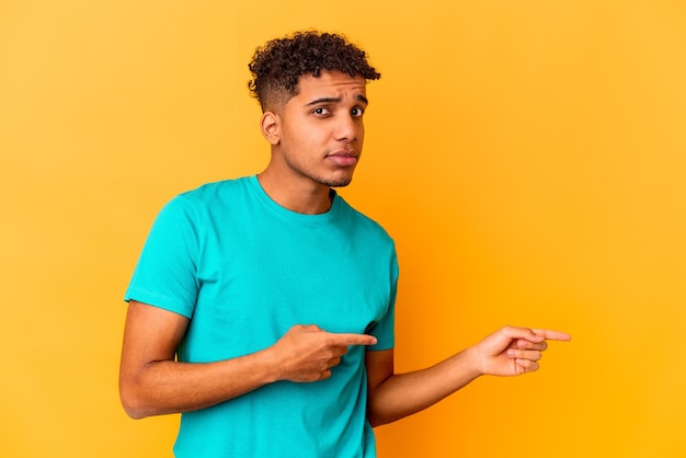 Joven afroamericano rizado en amarillo sorprendido apuntando con los dedos índices a un espacio de copia.