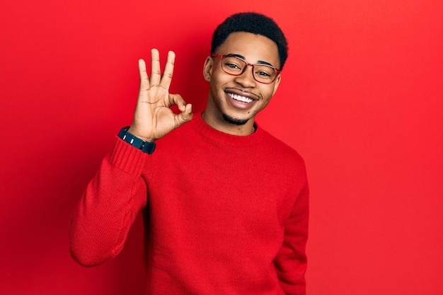 Joven afroamericano con ropa casual y anteojos sonriendo positivamente haciendo el signo de ok con la mano y los dedos expresión exitosa