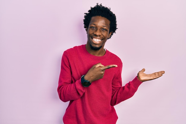 Joven afroamericano con ropa informal asombrado y sonriendo a la cámara mientras presenta con la mano y señala con el dedo.