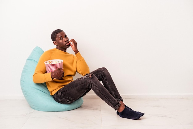 Joven afroamericano sentado en una bocanada comiendo palomitas de maíz aislado de fondo blanco mordiéndose las uñas nervioso y muy ansioso