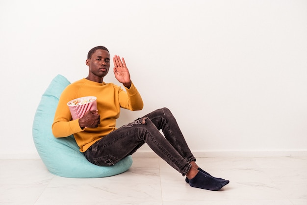 Joven afroamericano sentado en una bocanada comiendo palomitas de maíz aislado sobre fondo blanco de pie con la mano extendida mostrando la señal de stop, impidiéndote.