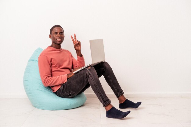 Joven afroamericano sentado en una bocanada usando una laptop aislada de fondo blanco que muestra el número dos con los dedos