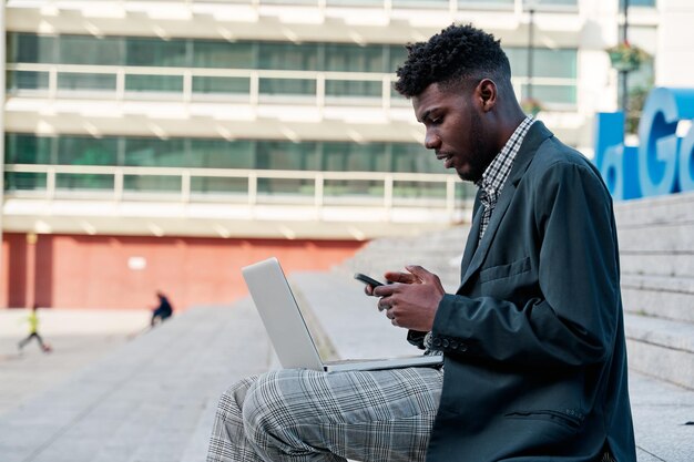 Joven afroamericano sentado en una escalera de la ciudad con su laptop mientras usa su teléfono móvil