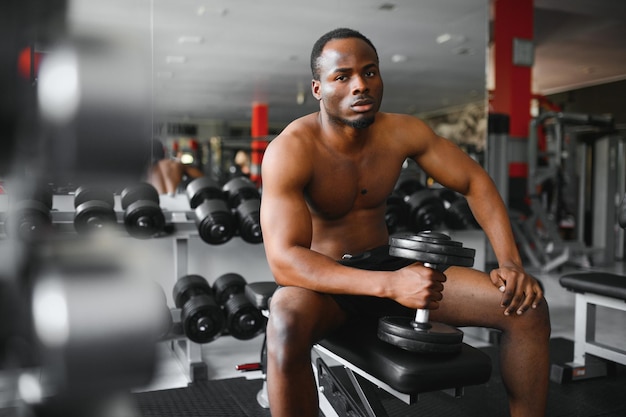 Joven afroamericano sentado y levantando una mancuerna cerca del estante en el gimnasio Persona de entrenamiento con pesas masculina haciendo un curl de bíceps en el gimnasio