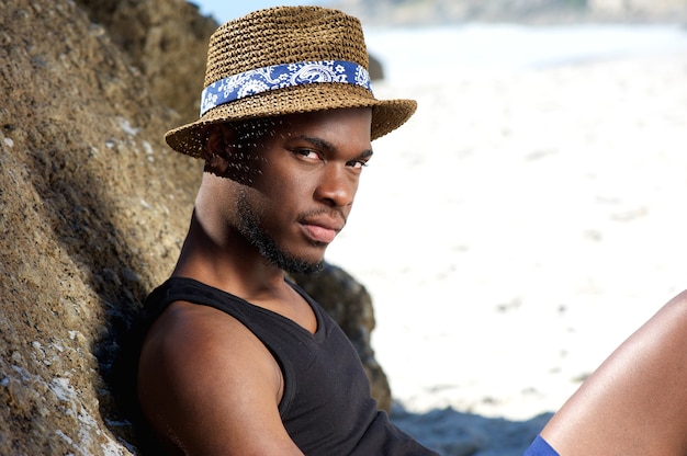 Joven afroamericano sentado en la playa con sombrero