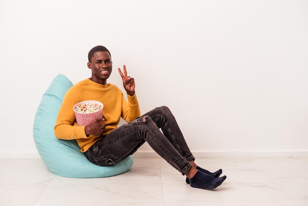 Joven afroamericano sentado en un soplo comiendo palomitas de maíz aislado en fondo blanco mostrando el número dos con los dedos.