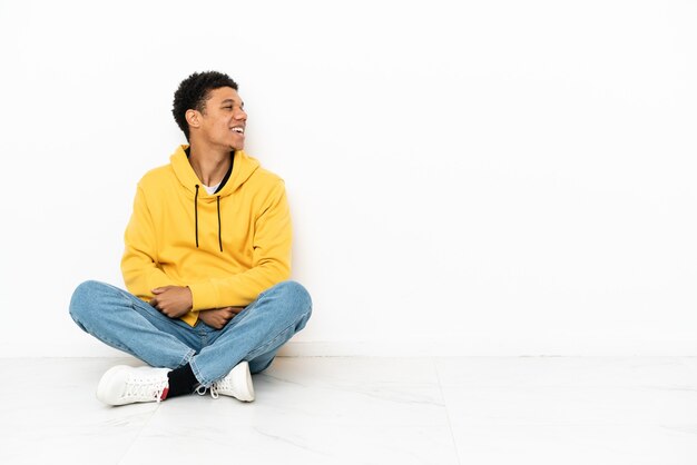 Joven afroamericano sentado en el suelo aislado sobre fondo blanco riendo en posición lateral