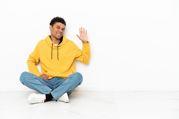 Joven afroamericano sentado en el suelo aislado sobre fondo blanco saludando con la mano con expresión feliz