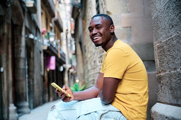 Foto joven afroamericano con smartphone y sonriendo en el casco antiguo de barcelona