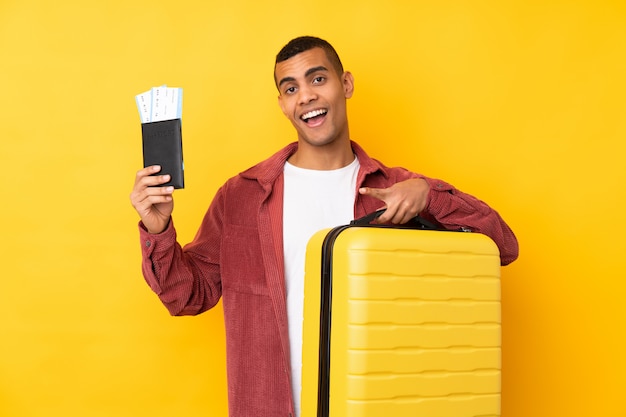 Joven afroamericano sobre pared amarilla aislada en vacaciones con maleta y pasaporte y sorprendido