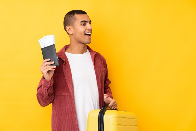 Joven afroamericano sobre pared amarilla aislada en vacaciones con maleta y pasaporte