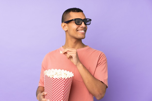 Joven afroamericano sobre pared azul aislada con gafas 3d y sosteniendo un gran cubo de palomitas de maíz