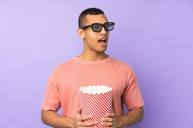 Joven afroamericano sobre pared azul aislado sorprendido con gafas 3d y sosteniendo un gran cubo de palomitas de maíz