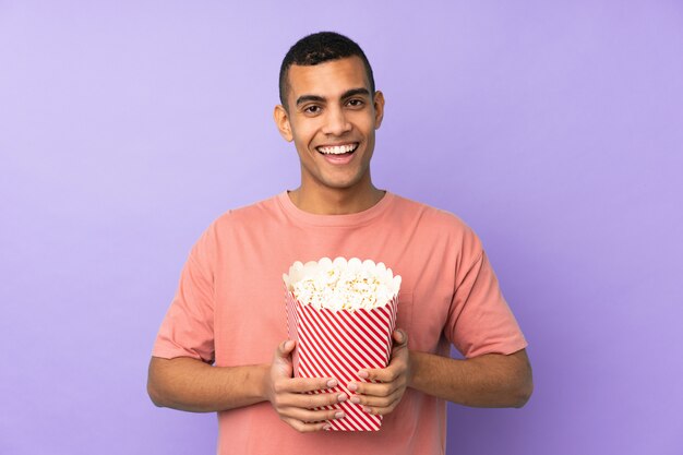 Joven afroamericano sobre pared azul aislado sosteniendo un gran cubo de palomitas de maíz