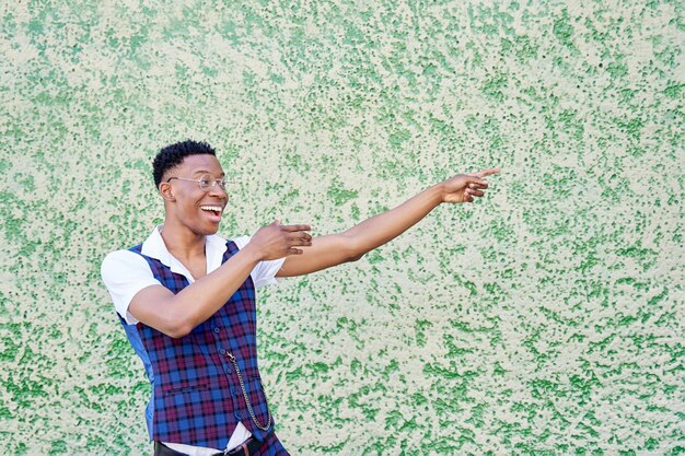 Joven afroamericano sonriendo alegremente sintiéndose feliz y apuntando hacia un lado y hacia arriba mostrando un objeto en el espacio de la copia contra la pared verde