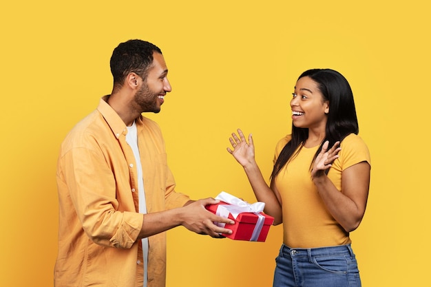 Un joven afroamericano sonriente le da una caja de regalo a una mujer sorprendida que felicita por el aniversario de vacaciones