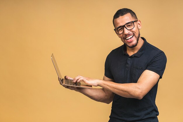 Joven afroamericano sonriente de pie y usando una computadora portátil aislada sobre un fondo beige