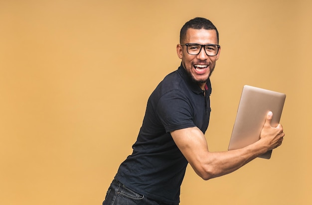 Joven afroamericano sonriente de pie y usando una computadora portátil aislada sobre un fondo beige