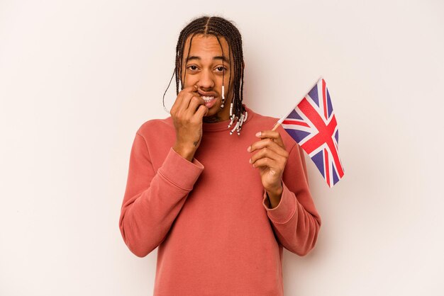 Joven afroamericano sosteniendo una bandera inglesa aislada de fondo blanco mordiéndose las uñas nervioso y muy ansioso