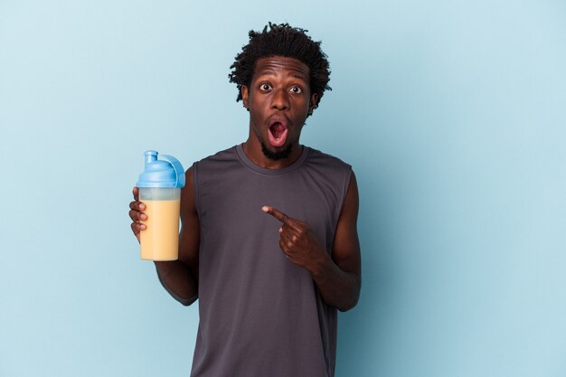 Joven afroamericano sosteniendo batido de proteínas aislado sobre fondo azul apuntando hacia el lado