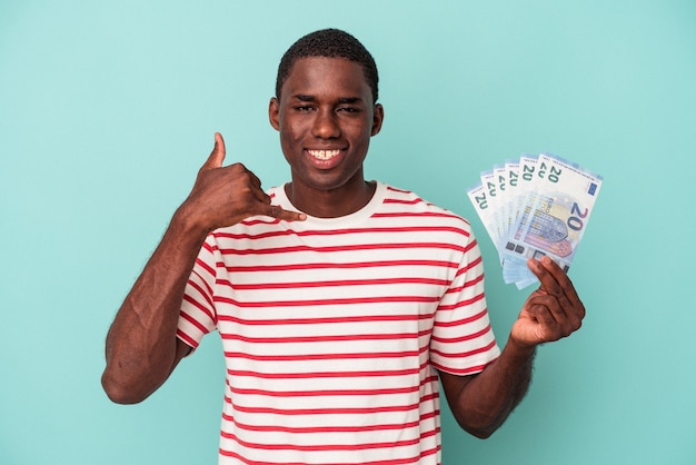 Joven afroamericano sosteniendo un billete de banco aislado sobre fondo azul que muestra un gesto de llamada de teléfono móvil con los dedos.