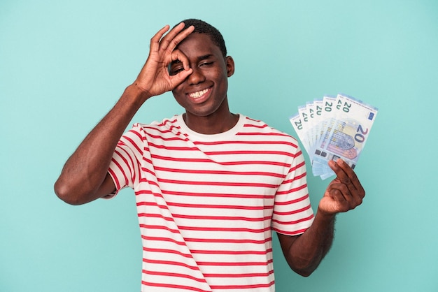 Joven afroamericano sosteniendo billetes de banco aislados en un fondo azul emocionado manteniendo el gesto correcto en el ojo