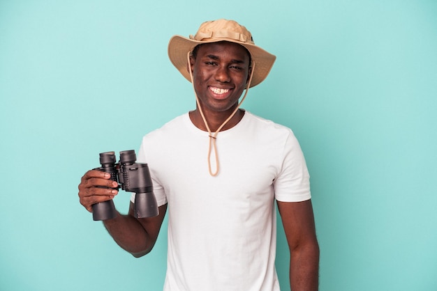 Foto joven afroamericano sosteniendo binoculares aislados de fondo azul feliz, sonriente y alegre.