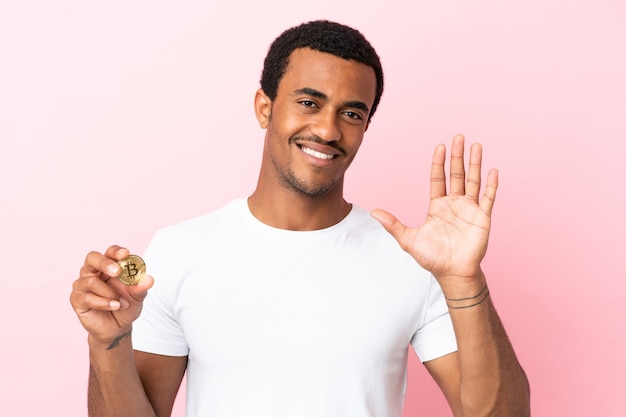Joven afroamericano sosteniendo un Bitcoin sobre fondo rosa aislado saludando con la mano con expresión feliz