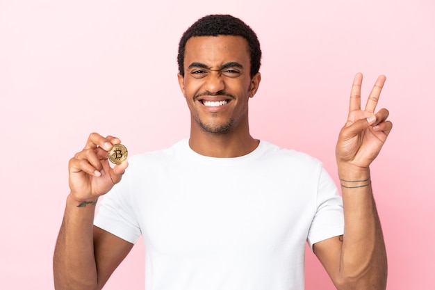 Joven afroamericano sosteniendo un Bitcoin sobre fondo rosa aislado sonriendo y mostrando el signo de la victoria