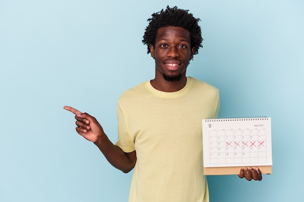 Joven afroamericano sosteniendo calendario aislado sobre fondo azul sonriendo y apuntando a un lado, mostrando algo en el espacio en blanco.