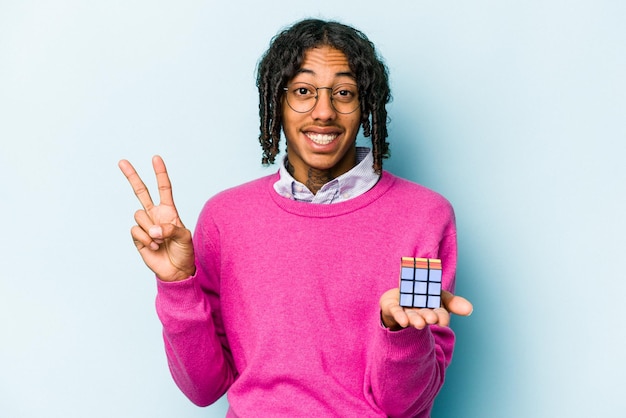 Joven afroamericano sosteniendo un cubo Rubiks aislado de fondo azul alegre y despreocupado mostrando un símbolo de paz con los dedos