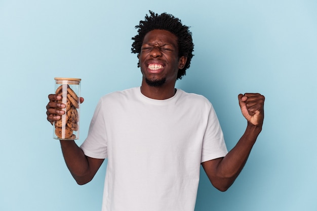 Joven afroamericano sosteniendo galletas de chispas de chocolate aislado sobre fondo azul.