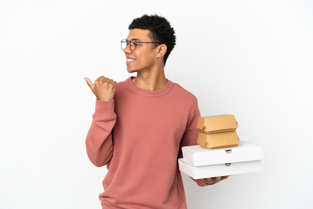 Joven afroamericano sosteniendo una hamburguesa y pizzas aislado sobre fondo blanco apuntando hacia el lado para presentar un producto