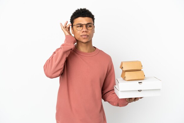 Joven afroamericano sosteniendo una hamburguesa y pizzas aislado sobre fondo blanco con dudas