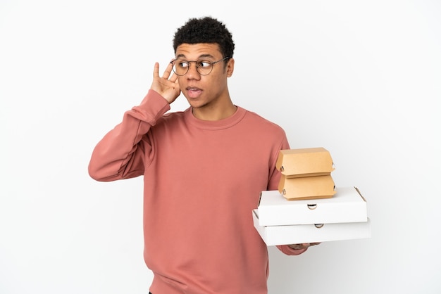 Joven afroamericano sosteniendo una hamburguesa y pizzas aislado sobre fondo blanco escuchando algo poniendo la mano en la oreja