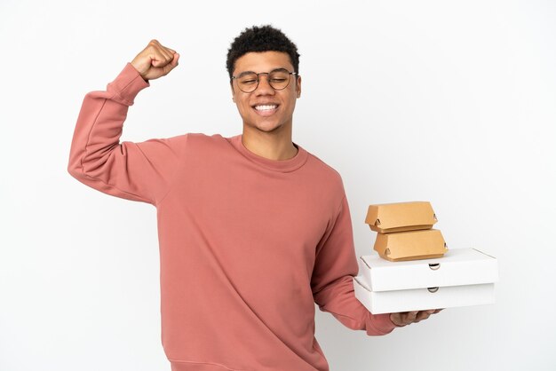 Foto joven afroamericano sosteniendo una hamburguesa y pizzas aislado sobre fondo blanco haciendo gesto fuerte