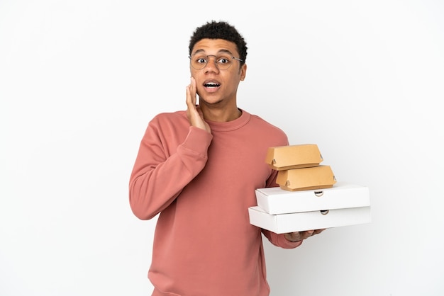 Joven afroamericano sosteniendo una hamburguesa y pizzas aislado sobre fondo blanco con sorpresa y expresión facial conmocionada