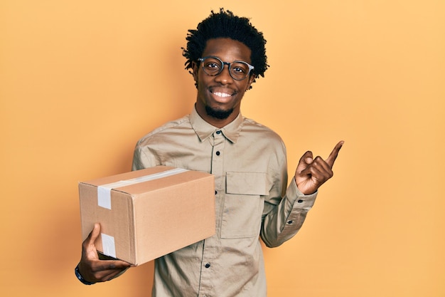 Joven afroamericano sosteniendo un paquete de entrega sonriendo feliz señalando con la mano y el dedo a un lado