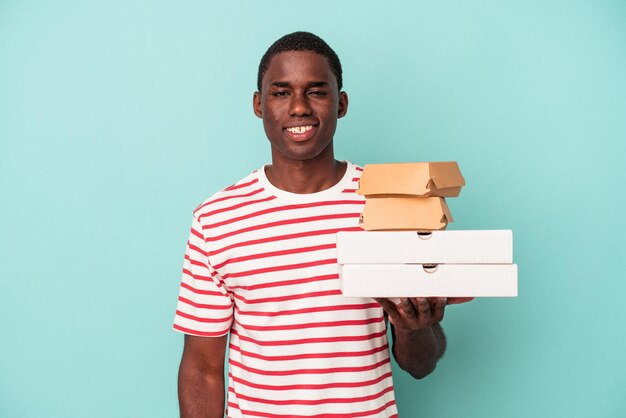 Joven afroamericano sosteniendo pizzas y hamburguesas aisladas de fondo azul feliz, sonriente y alegre.