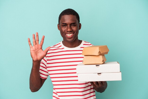 Joven afroamericano sosteniendo pizzas y hamburguesas aisladas de fondo azul sonriendo alegremente mostrando el número cinco con los dedos.