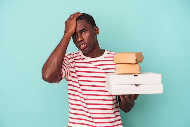 Joven afroamericano sosteniendo pizzas y hamburguesas aisladas sobre fondo azul conmocionado, ella ha recordado una reunión importante.