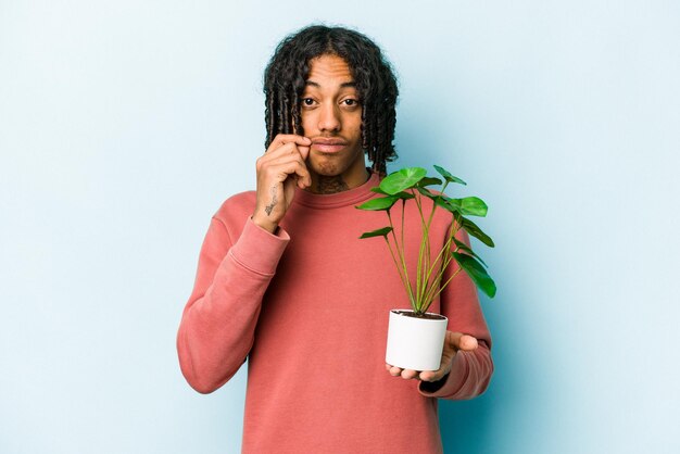 Joven afroamericano sosteniendo una planta aislada de fondo azul con los dedos en los labios guardando un secreto