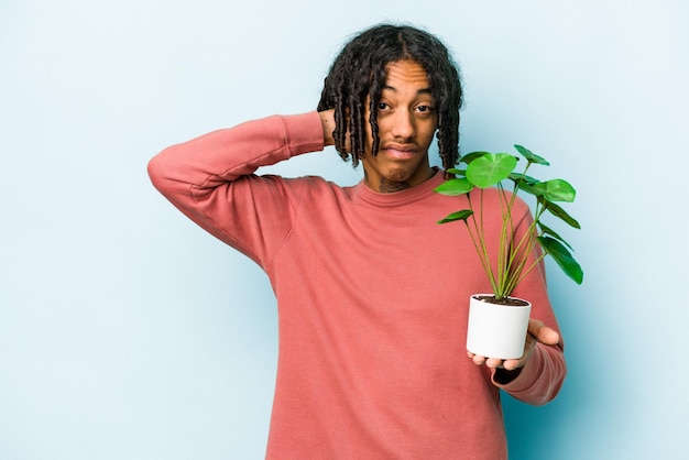 Joven afroamericano sosteniendo una planta aislada de fondo azul tocando la parte posterior de la cabeza pensando y tomando una decisión