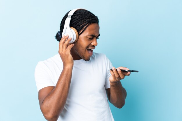 Joven afroamericano con trenzas aislado sobre fondo azul escuchando música con un móvil y cantando