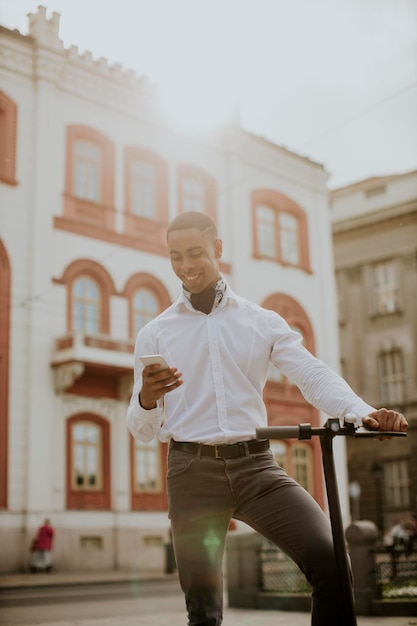 Joven afroamericano usando teléfono móvil mientras está de pie con scooter eléctrico en una calle