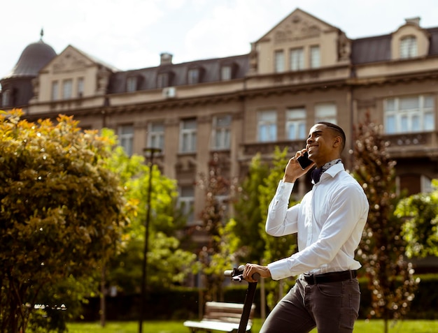 Joven afroamericano usando teléfono móvil mientras está de pie con scooter eléctrico en una calle