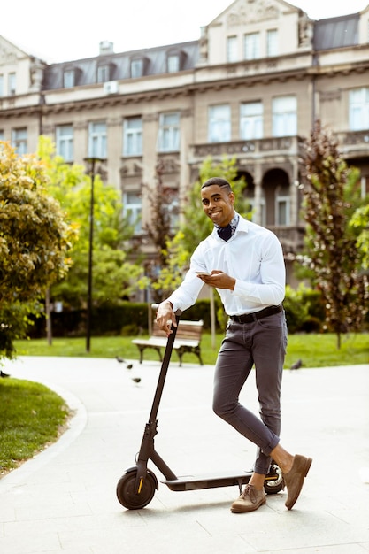 Joven afroamericano usando teléfono móvil mientras está de pie con scooter eléctrico en una calle