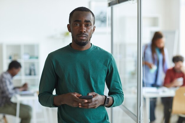 Joven afroamericano vestido con ropa casual y sosteniendo el teléfono inteligente mientras está de pie en la oficina